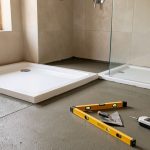 Chape fraîchement nivelée dans une salle de bains, avec un receveur de douche blanc et des outils de contrôle disposés à proximité.
